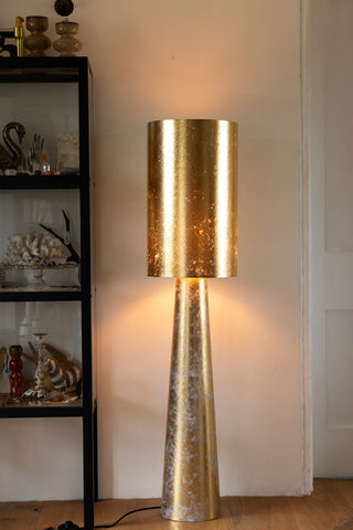 The Gold Leafed Effect Floor Lamp switched on in front of a white wall next to a black and glass display cabinet.