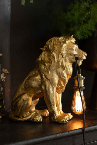 The Quirky Gold Lion Lamp switched on and displayed on a black sideboard with some greenery.