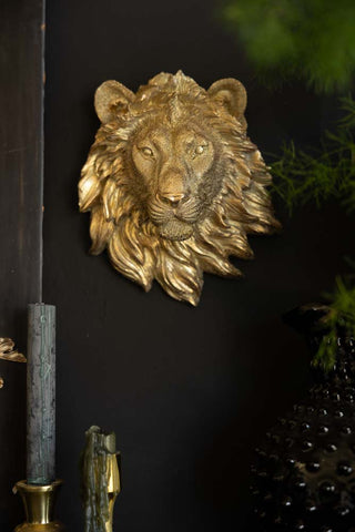 A gold lion head ornament on a black wall, styled with candles, greenery and a jug.