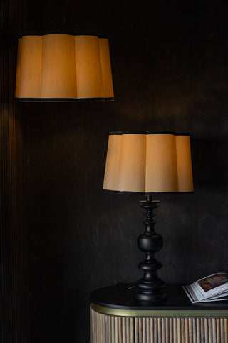 Two Gold Scalloped Easyfit Lampshades, one styled on a lamp base next to an open book on a wooden unit and the other styled as a pendant light in front of a dark wall, both with lights switched on.