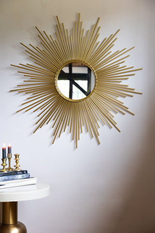 A round mirror with starburst design styled on a white wall, above a table with lit candles and books on.