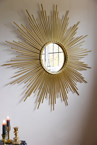 A round mirror with starburst design, styled on a white wall above some lit candles in candlestick holders.