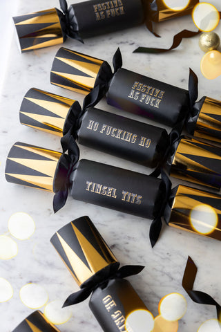 Set of 6 black and gold sweary crackers laid out on a table with some festive sparkle