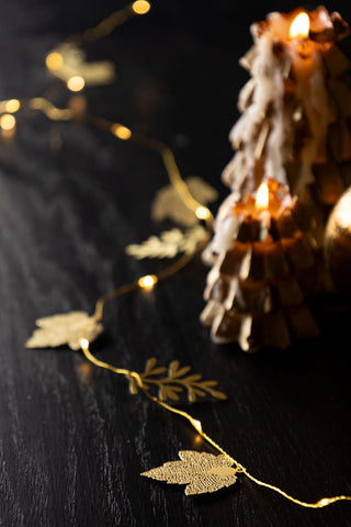 Two christmas tree candles burining in the background, while the golden leaves light chain is decorated on the table around them.