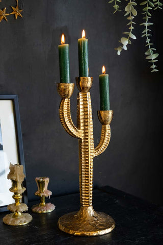 The Gorgeous Gold Cactus Candle Holder styled with lit candles inside, with other candlestick holders, greenery, a star ornament and a frame in the background.