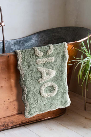 The Green Ciao Bath Mat draped across the edge of a copper bath, styled with a plant.