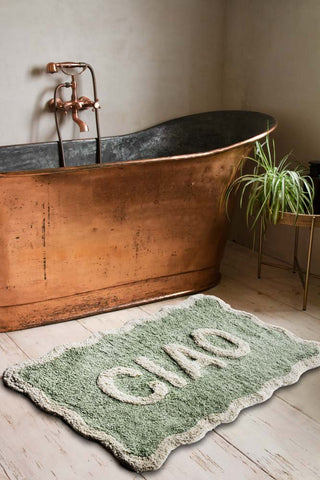 The Green Ciao Bath Mat styled next to a copper bath, with a side table and plant.