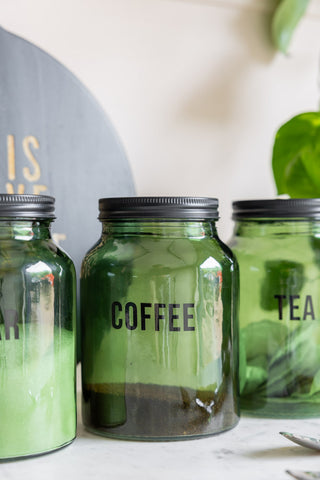 The Green Glass Storage Jar With Black Lid - Coffee on a white marble counter with the two matching jars, a serving board and a plant.