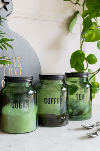 The Green Glass Storage Jar With Black Lid - Sugar with the matching tea and coffee jars, styled on a marble counter with other kitchen accessories and plants.