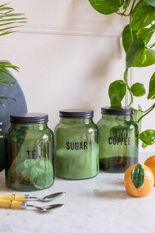 The Green Glass Storage Jar With Black Lid - Sugar displayed with the matching tea and coffee versions, styled with various kitchen accessories and plants.