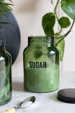 The Green Glass Storage Jar With Black Lid - Sugar with sugar inside, styled with other kitchen accessories and plants.