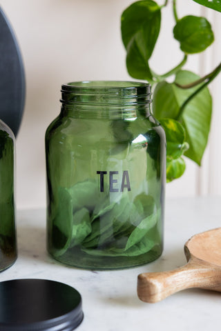 The Green Glass Storage Jar With Black Lid - Tea with the lid off and teabags inside, styled with other kitchen accessories and a plant.