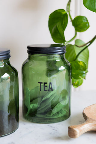 The Green Glass Storage Jar With Black Lid - Tea styled with teabags inside on a marble surface, with other kitchen accessories and a plant.