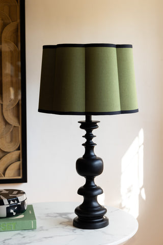 The Green Scalloped Easyfit Lampshade styled on a lamp base on a white marble table, with a book, trinket box and piece of artwork.