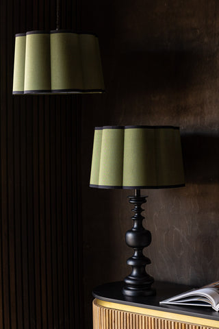 Two Green Scalloped Easyfit Lampshades, one styled on a black lamp base on a unit with an open book, and the other hanging as a pendant light in front of a dark wall.