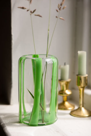 The Green Striped Glass Vase displayed on a window ledge with some flower stems inside, next to two gold candlestick holders.