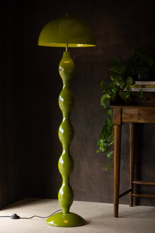 The Green Wavy Retro Floor Light switched on and styled in front of a dark wall and a desk styled with some books and a plant.