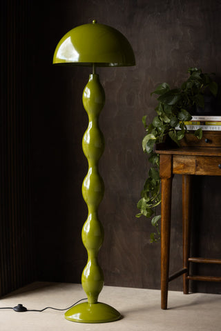 A green wavy floor lamp styled in a dark room with a desk, books and greenery.