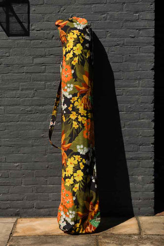 The HKliving Dark Florals Flourish Parasol in its bag, leaning against a black brick wall.