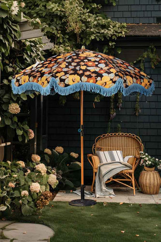 The HKliving Floral Flourish Parasol with Blue Fringing styled in a garden with a wicker chair and plants.