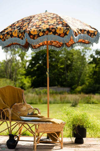 The HKliving Floral Flourish Parasol with Blue Fringing in a garden in the sun, styled with a wicker lounge chair.