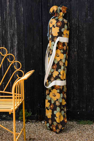 The HKliving Floral Flourish Parasol with Blue Fringing in its bag, leaning against a black fence next to a bench.