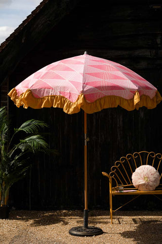The HKliving Pink Linear Parasol with Mustard Fringe in the sunshine, styled next to a yellow bench with cushion and a plant.