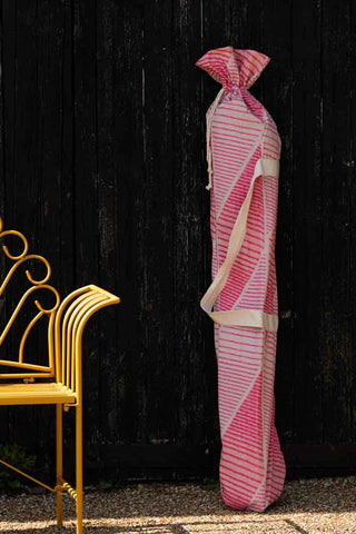 The HKliving Pink Linear Parasol with Mustard Fringe in its bag, leaning against a black fence next to a yellow bench.