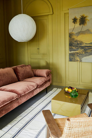 The HKliving Retro Sofa in Magnolia Velvet seen from a side angle, styled in a living room with a large pendant light, coffee table, rug and chair, styled with some decorative accessories.