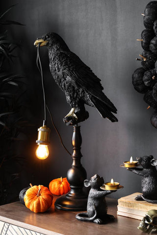 Black crow lamp on a sideboard styled with orange velvet mini pumpkins, mice candles and a skull ornament. The crow lamp is lit and the setting is giving Halloween vibes.