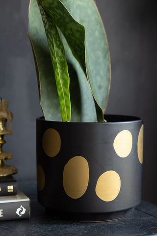 The Handcrafted Black & Metallic Planter in Medium styled on a black sideboard with some books and a candlestick holder, with a plant inside.