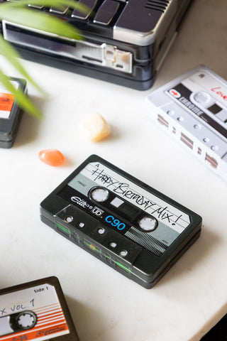 The Happy Birthday Cassette Tape Storage Tin with the lid on, styled on a white marble surface with various accessories and other similar storage tins.