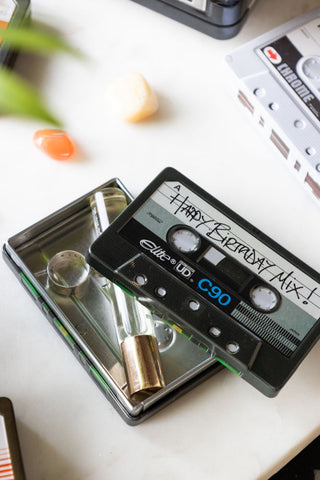 The Happy Birthday Cassette Tape Storage Tin with the lid off, styled with some accessories inside and displayed on a surface with other storage tins.