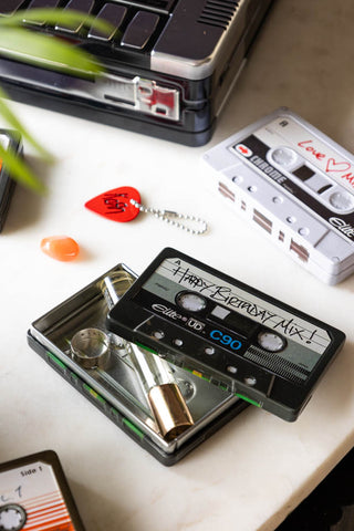 The Happy Birthday Cassette Tape Storage Tin styled on a marble surface with various accessories, with other storage tins and a plant.