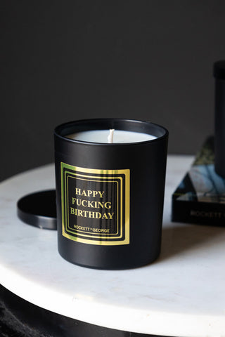 The Happy Fucking Birthday Scented Candle with the lid off, displayed on a white table with a book.