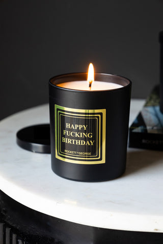 The Happy Fucking Birthday Scented Candle lit and displayed on a white surface, with the lid and a book in the background.