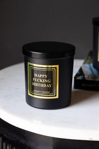 The Happy Fucking Birthday Scented Candle with the lid on, displayed on a white surface with a book.