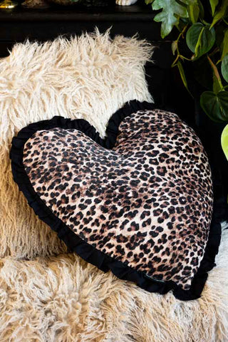 The Heart Leopard Print Velvet Ruffle Cushion styled on a cream fur throw in front of a black cabinet.