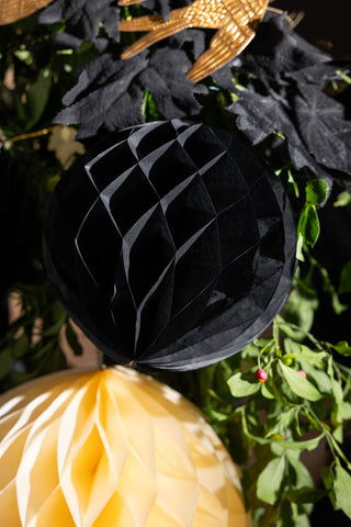 The Set Of 2 Black Honeycomb Ball Decorations styled in front of a green leaf garland and next to a matching cream paper decoration.