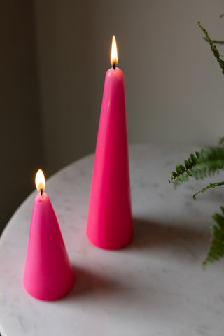 The Small and Large Hot Pink Cone Shaped Candles displayed together on a white marble table lit, with a plant to the side of the shot.