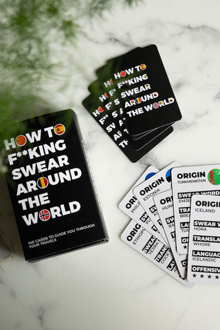 The How To Swear Around the World Card Pack displayed on a marble table with some greenery.