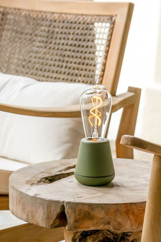 The Humble - Outdoor Cable-Free Mood Lighting in Moss Green displayed on a wooden side table, next to some chairs.