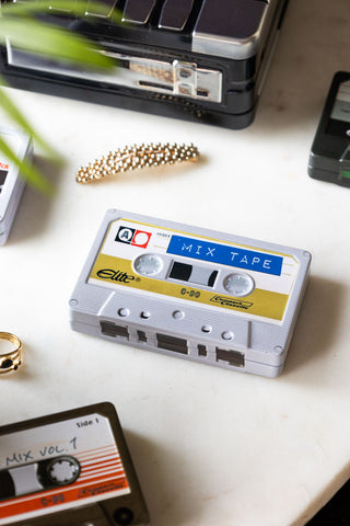 The I Made You A Mix Tape Cassette Tape Storage Tin with the lid on, styled on a white surface with a clip and a ring, displayed with other storage tins and a plant.