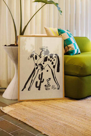 A graphic and fun art print framed and leaning against a green sofa. The art print features a cowgirl on a horse with catci underneath.