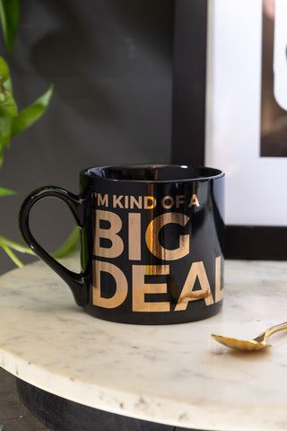 The I'm Kind Of A Big Deal Mug displayed on a marble surface with a spoon and framed artwork, with the leaves of a plant in the background.