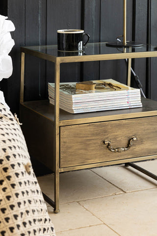 An industrial style bedside table with some magazines, a mug and a lamp on, displayed next to a bed.