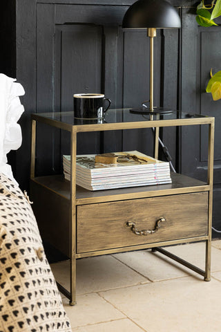 An industrial style bedside table styled next to a bed, with a stack of magazines, a mug and a table lamp.