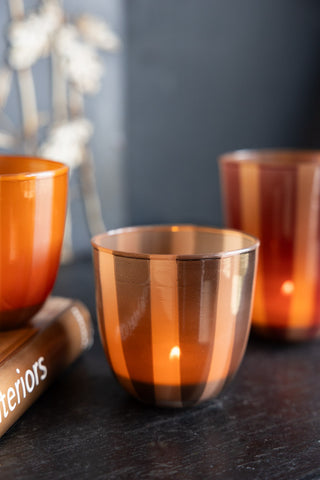 The Khaki Striped Tealight Holder - Short with a lit tealight inside, displayed on a black surface with a book and two other tealight holders.