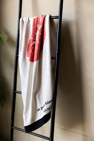 The Kiss Me Beach Towel hanging on a black rail in front of a neutral wall, with a plant in the background.