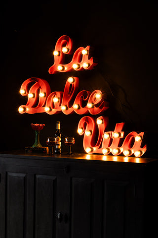 The La Dolce Vita Fairground Wall Light displayed illuminated on the wall above a flower base, a bottle and some wine glasses.